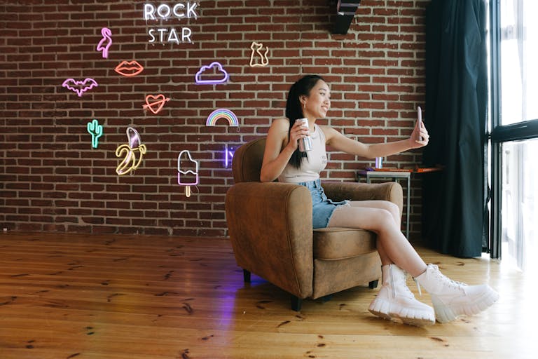 Young woman in stylish casual wear taking a selfie in a chic room with neon decor and brick walls.