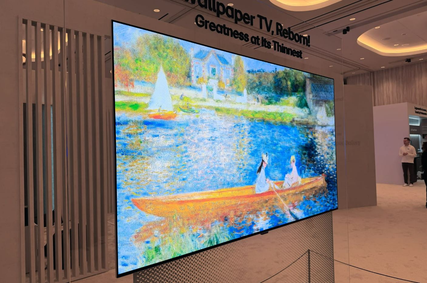 A imagem apresenta uma televisão OLED de última geração da LG, destacada pela sua espessura extremamente reduzida, instalada num ambiente de exposição moderno e minimalista. O ecrã, que parece flutuar junto à parede devido ao seu design "Wallpaper", exibe uma pintura impressionista vibrante que retrata uma cena ribeirinha com barcos à vela e figuras humanas numa embarcação a remos, utilizando cores vivas como azuis, amarelos e verdes. Acima da televisão, na parede branca, lê-se a inscrição "Wallpaper TV, Reborn! Greatness at Its Thinnest", reforçando o conceito de tecnologia wireless e ultra-fina. O televisor está apoiado numa base metálica com um padrão perfurado, situada num espaço amplo com iluminação embutida no teto e tons claros. Ao fundo, no lado direito, vislumbra-se a silhueta de uma pessoa num balcão de atendimento, inserida num contexto que sugere ser uma feira tecnológica ou um showroom de eletrónica de consumo.