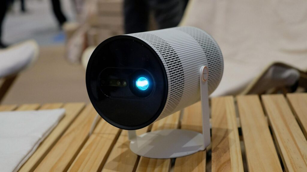 A imagem apresenta o projetor portátil Samsung The Freestyle, um dispositivo com um design moderno e cilíndrico em tons de branco e preto. O projetor está montado num suporte giratório que permite ajustar facilmente o ângulo de projeção, encontrando-se pousado sobre uma mesa de ripas de madeira clara, num ambiente que sugere ser uma feira tecnológica ou um espaço de demonstração. A parte frontal é composta por uma face preta circular onde se destaca a lente principal, que emite uma luz azul brilhante, ladeada por sensores de focagem e nivelamento automático. O corpo do dispositivo possui uma textura perfurada, ideal para a dissipação de calor e para a saída de som omnidirecional. Na lateral, é visível o mecanismo de dobradiça com um detalhe em laranja, que confere um toque de cor ao design minimalista. O fundo da imagem está ligeiramente desfocado, mostrando figuras humanas e mobiliário de tons neutros, o que mantém o foco visual inteiramente no projetor.