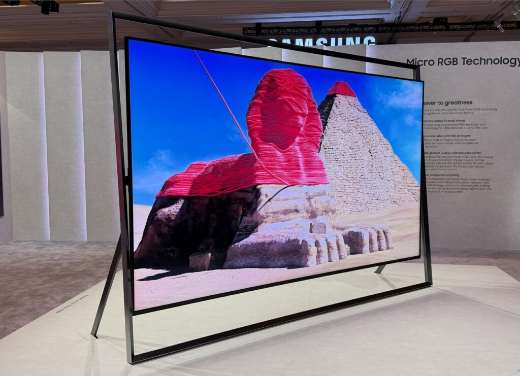 A imagem apresenta uma televisão de grandes dimensões da Samsung, destacando-se como o modelo de topo com tecnologia Micro RGB, exposta num evento que parece ser a CES 2026. O ecrã exibe uma imagem vibrante e detalhada da Esfinge e das Pirâmides de Gizé, no Egito, onde a Esfinge surge coberta por uma textura vermelha em camadas, criando um contraste acentuado com o céu azul e os tons arenosos do deserto. A estrutura do televisor é minimalista e moderna, composta por uma moldura fina e preta que se estende para além dos limites do ecrã, formando um suporte em estilo de cavalete que assenta diretamente no chão. No fundo, a parede branca do pavilhão exibe o logótipo da Samsung e o texto informativo sobre a tecnologia Micro RGB, explicando as vantagens do novo motor de inteligência artificial e a precisão das cores. O ambiente é iluminado de forma suave, típico de uma feira tecnológica, focando todas as atenções na clareza e no brilho do ecrã.