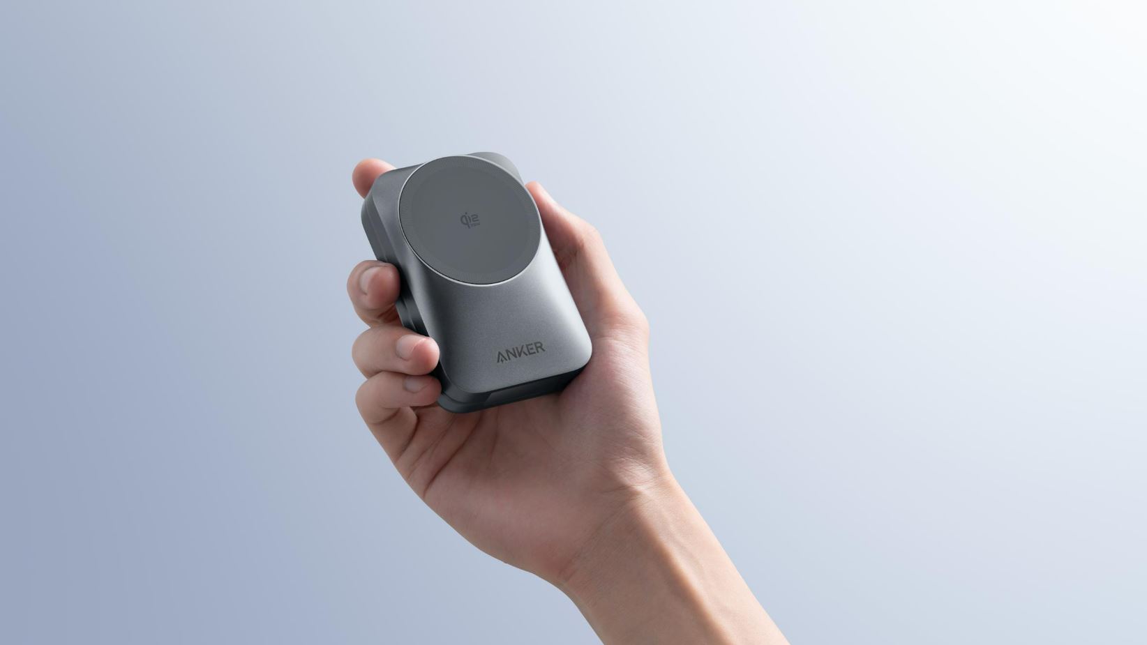 Esta imagem promocional foca-se num novo dispositivo de carregamento portátil da marca Anker, que ostenta a certificação Qi2. O objeto é segurado por uma mão humana, destacando o seu design compacto e ergonómico, com dimensões que parecem ideais para caber facilmente na palma da mão ou num bolso. O carregador apresenta um acabamento num tom cinza-escuro metalizado (Space Gray), com cantos arredondados que lhe conferem um aspeto moderno e robusto. Na parte frontal, destaca-se um círculo ligeiramente elevado com uma textura diferente, onde se lê o logótipo "Qi2 15W", indicando a sua compatibilidade com carregamento magnético de alta velocidade. Abaixo desta zona de contacto, o nome da marca "ANKER" surge gravado de forma discreta. carregador Anker qi2 apresentado na CES 2026