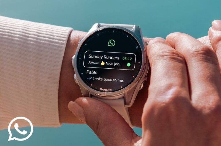 A imagem apresenta um plano aproximado do pulso de uma pessoa a utilizar um smartwatch prateado da marca Garmin, com uma bracelete branca de material suave. O foco central está no mostrador circular do relógio, que exibe a interface da aplicação WhatsApp. No ecrã, é visível o logótipo verde da app no topo e uma lista de notificações de mensagens: destaca-se uma mensagem do grupo "Sunday Runners", onde um contacto chamado Jordan enviou um emoji de polegar para cima acompanhado do texto "Nice job!", e logo abaixo surge uma conversa individual com "Pablo", que escreveu "Looks good to me". A mão da pessoa, com unhas cuidadas, toca suavemente na lateral da caixa do relógio, sugerindo a interação com os botões físicos do dispositivo. O fundo da imagem está desfocado, revelando tons quentes de pele e um ambiente que parece ser ao ar livre, com uma luz solar suave que realça os detalhes metálicos do relógio. No canto inferior esquerdo da composição, está sobreposto o logótipo oficial do WhatsApp em branco, reforçando o tema da conectividade e da integração da aplicação no ecossistema Garmin.