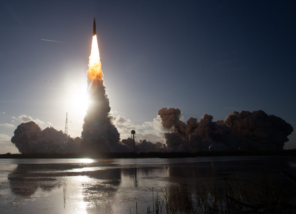 Foguetão SLS da missão Artemis II em plena descolagem, com uma coluna de fogo brilhante e densas nuvens de fumo refletidas na água em primeiro plano.