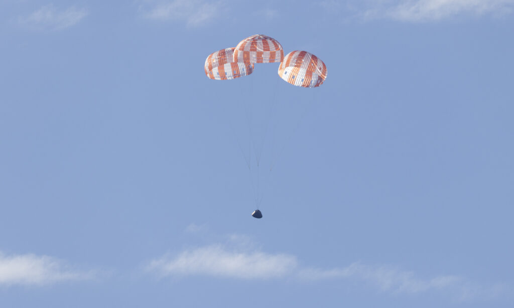 Cápsula espacial Orion a descer suavemente sob três paraquedas cor de laranja e brancos num céu azul límpido.