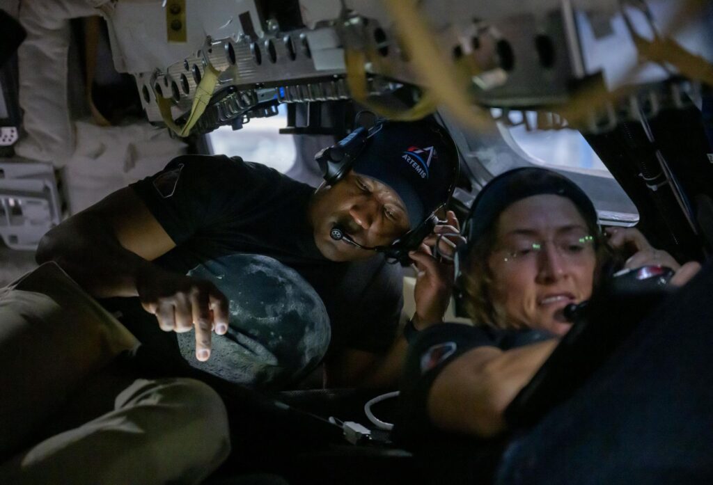 Victor Glover e Christina Koch a trabalhar dentro de um simulador de cápsula espacial, rodeados por estruturas metálicas e utilizando auscultadores de comunicação.