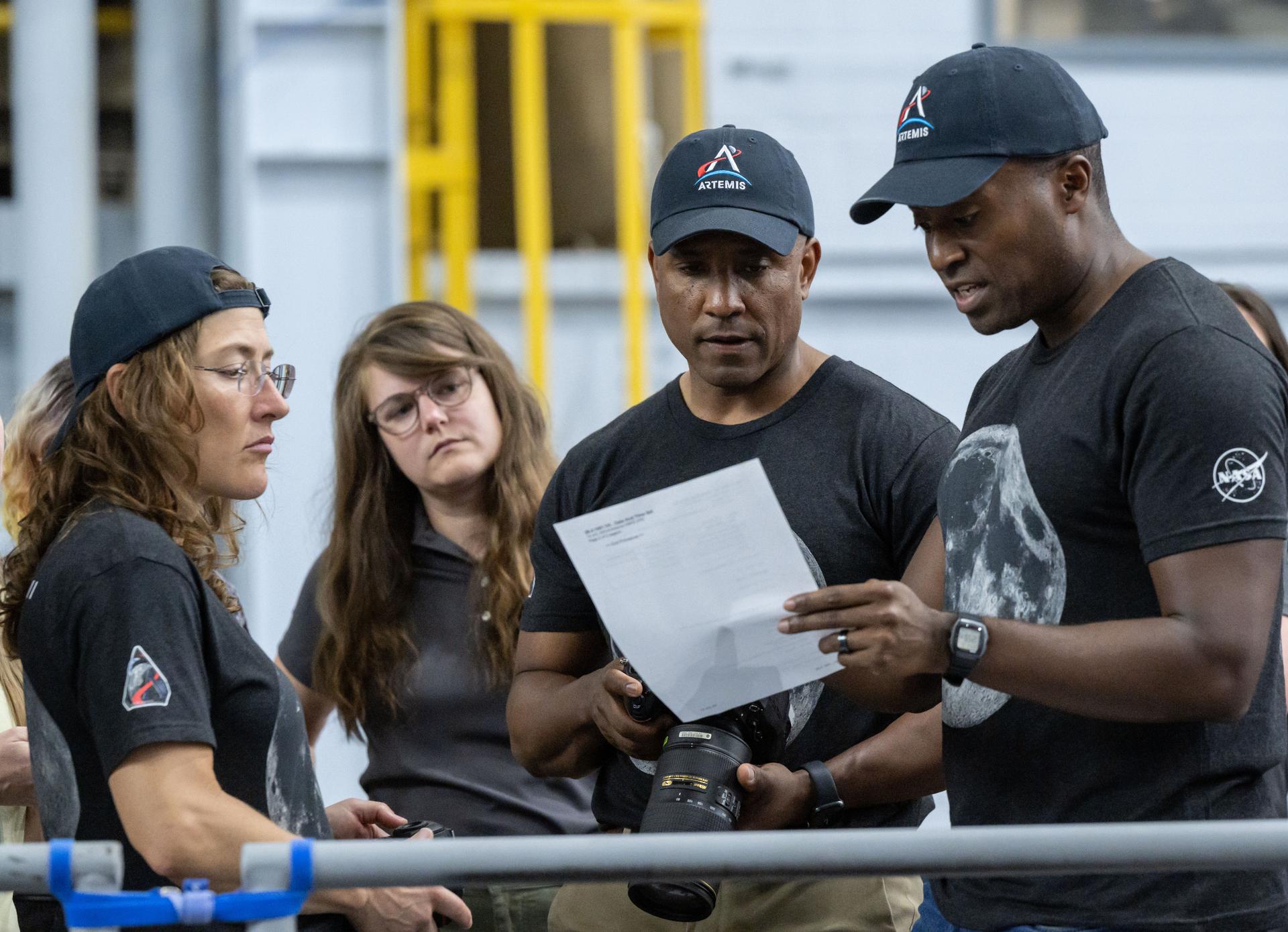 Christina Koch, Marie Henderson e Victor Glover, vestidos com t-shirts da Artemis, analisam um documento impresso e seguram uma câmara fotográfica num hangar de treino.
