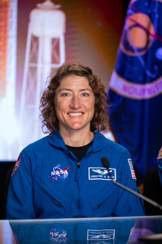 Retrato da astronauta Christina Hammock Koch, especialista de missão da Artemis II, sorrindo e vestida com o seu fato de voo azul da NASA. O uniforme mostra o logótipo da NASA e um crachá com o seu nome. Ela está sentada atrás de uma mesa transparente, com um fundo desfocado que exibe uma torre de lançamento e uma bandeira oficial.