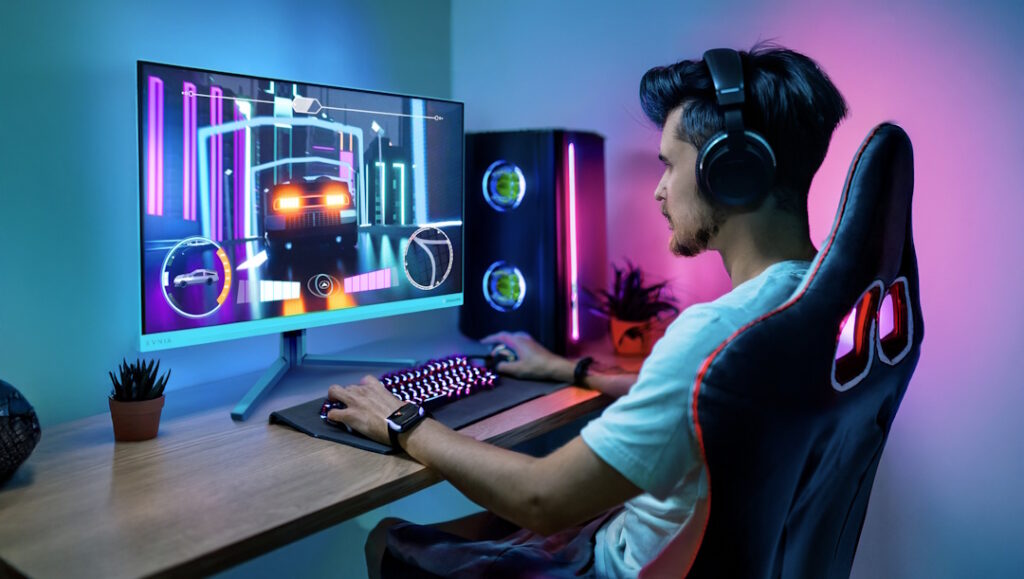 Fotografia de um jogador com auscultadores sentado numa cadeira de gaming, focado a jogar um jogo de corridas num monitor Philips Evnia. O ambiente é iluminado por luzes vibrantes em tons de azul à esquerda e rosa à direita. Na secretária de madeira, o jogador usa um teclado RGB e um rato, tendo ao fundo uma torre de computador com iluminação interna e um pequeno vaso com uma planta.
