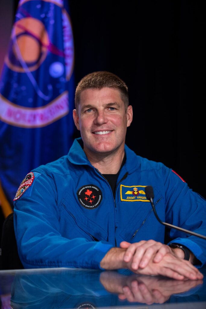 Retrato do astronauta canadiano Jeremy Hansen, especialista de missão da Artemis II, vestido com o seu fato de voo azul. O uniforme exibe o logótipo da Agência Espacial Canadiana e um crachá amarelo com o seu nome. Ele está com as mãos cruzadas sobre uma mesa transparente, com uma bandeira oficial desfocada ao fundo.