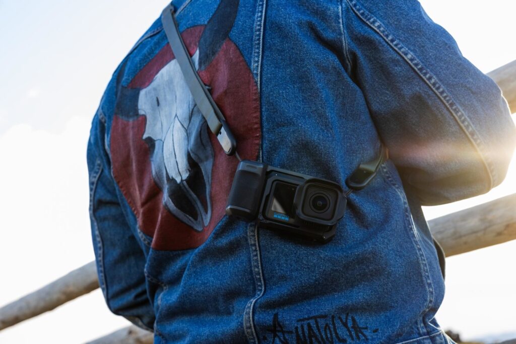 Vista traseira de uma pessoa usando uma jaqueta jeans azul com uma grande estampa de caveira de búfalo em um círculo vermelho. Uma câmera de ação GoPro preta está pendurada diagonalmente sobre o ombro da pessoa por uma alça tática. A luz do sol brilha intensamente no lado direito da imagem.