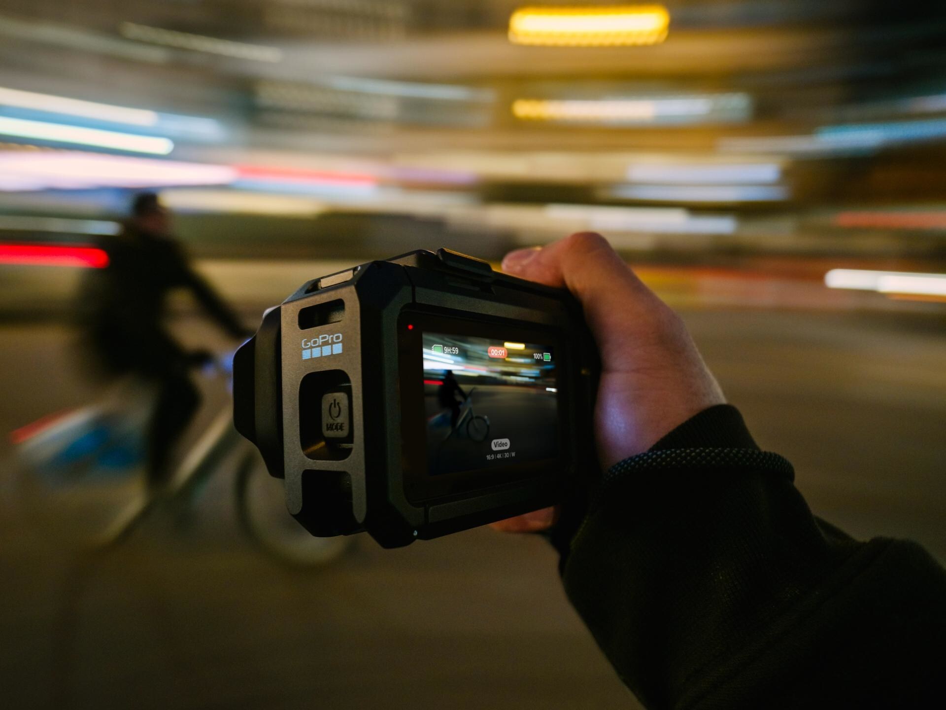 Uma mão segura firmemente uma câmara de ação GoPro preta e protegida, em primeiro plano, com o ecrã ativado a gravar, contra um fundo noturno desfocado e em movimento que mostra rastos de luz e um ciclista a passar rapidamente. No corpo da câmara, lê-se "GOPRO" e o botão "MODE".