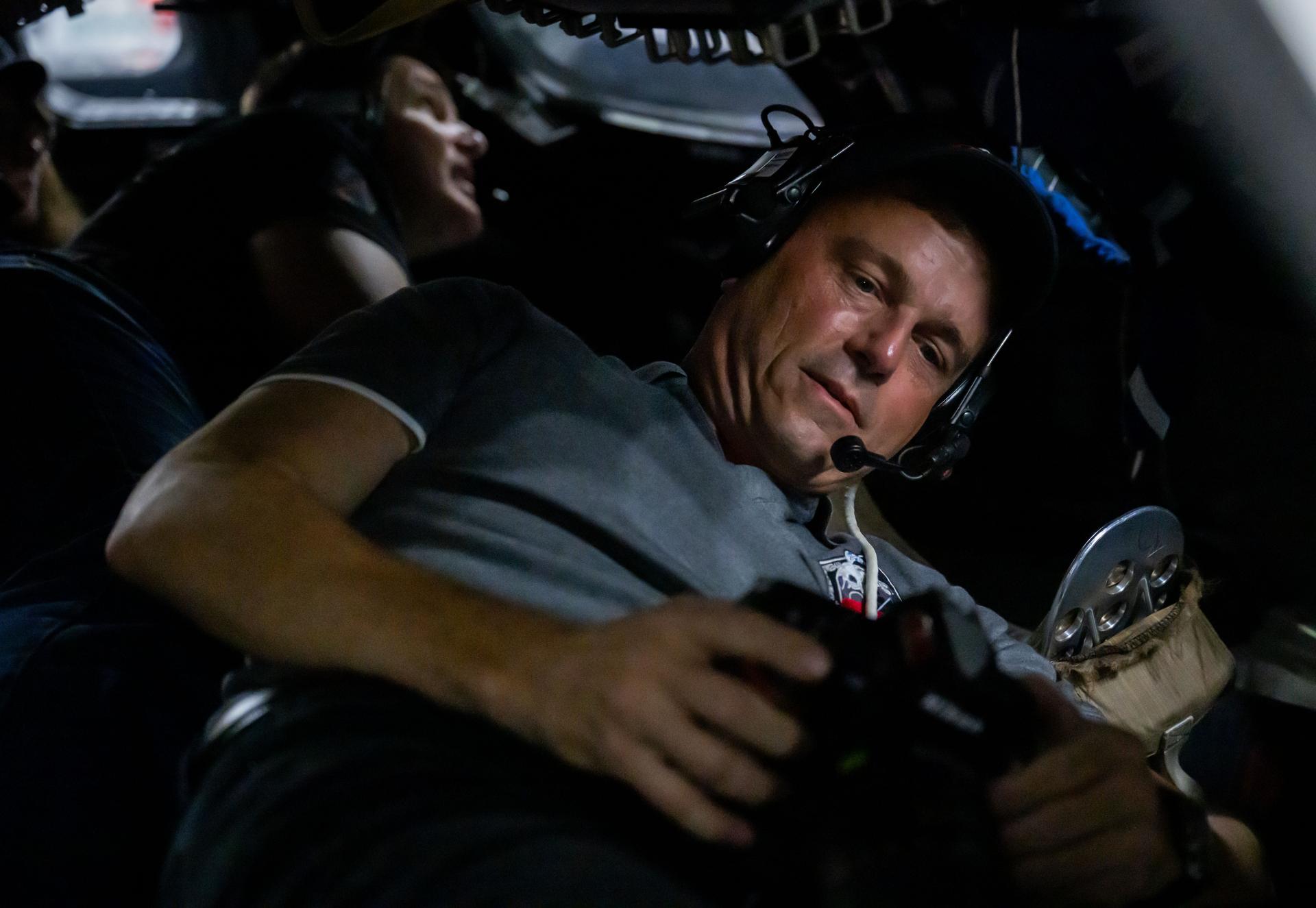 Grande plano do astronauta Reid Wiseman com auscultadores de comunicação, operando controlos num ambiente de simulação de cabine espacial com iluminação ténue.