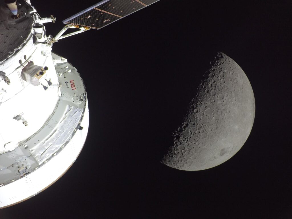 Parte da cápsula espacial Orion com logótipos da NASA e ESA em órbita, com a superfície craterada da Lua visível ao fundo.