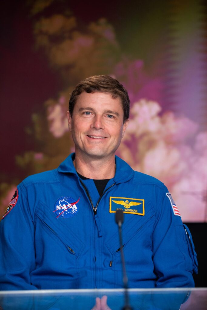 Retrato de meio corpo do astronauta Reid Wiseman, comandante da missão Artemis II, sorrindo e vestido com o seu fato de voo azul oficial da NASA. O fato exibe o logótipo da NASA, a bandeira americana e um crachá de identificação amarelo. Wiseman está sentado atrás de uma mesa transparente, com um fundo desfocado em tons de rosa e roxo.