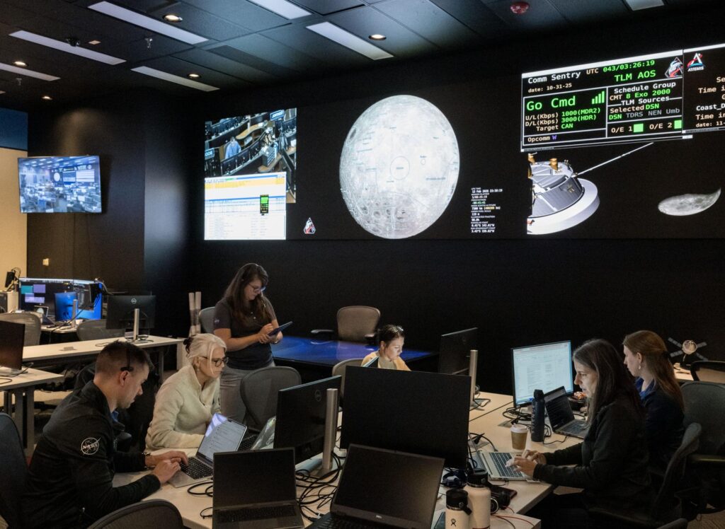 Equipa da missão Artemis II a trabalhar em computadores numa sala de controlo escura, com um grande ecrã ao fundo exibindo a face da Lua e dados técnicos de telemetria.