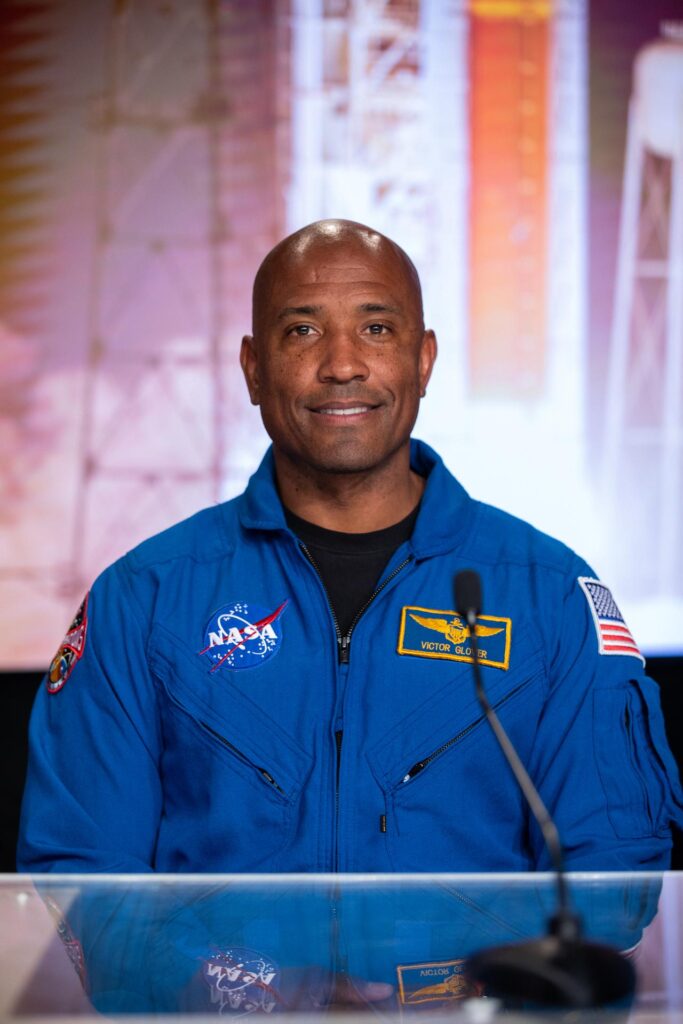 Retrato do astronauta Victor Glover, piloto da missão Artemis II, vestido com o fato de voo azul da NASA. O uniforme inclui o logótipo da NASA, a bandeira americana e um crachá amarelo com o seu nome. Ele está sentado atrás de uma mesa de vidro, com um fundo desfocado que mostra um foguetão na plataforma de lançamento.
