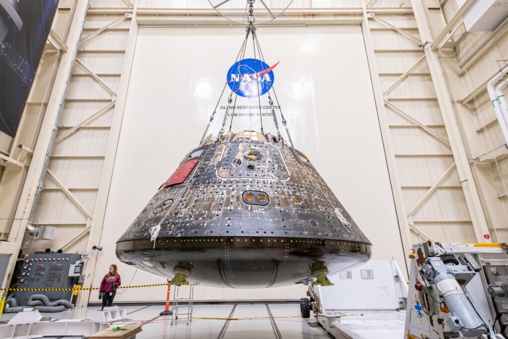 No interior de um grande hangar no Glenn Research Center da NASA, o módulo de tripulação da Orion, com um aspeto escuro e bronzeado do escudo térmico e painéis técnicos expostos, está suspenso por cabos de aço no centro. No fundo, a parede branca ostenta um grande logótipo da NASA e o texto "GLENN RESEARCH CENTER PLUM BROOK STATION". Uma técnica monitoriza a operação a partir do chão, ao lado de equipamento de suporte industrial.