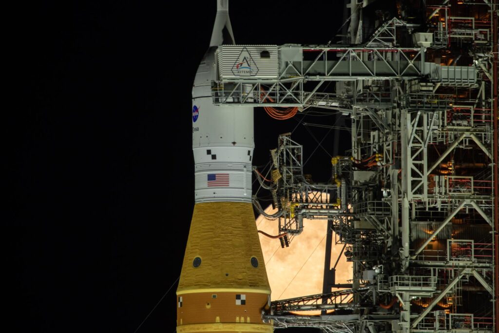 Uma fotografia noturna e dramática da silhueta do foguetão SLS e da cápsula Orion na plataforma de lançamento. A nave espacial é emoldurada contra o disco gigante e detalhado de uma lua cheia amarelada no céu preto. A estrutura metálica da torre de lançamento rodeia o veículo lunar, criando um contraste visual impressionante entre a engenharia humana e o satélite natural.