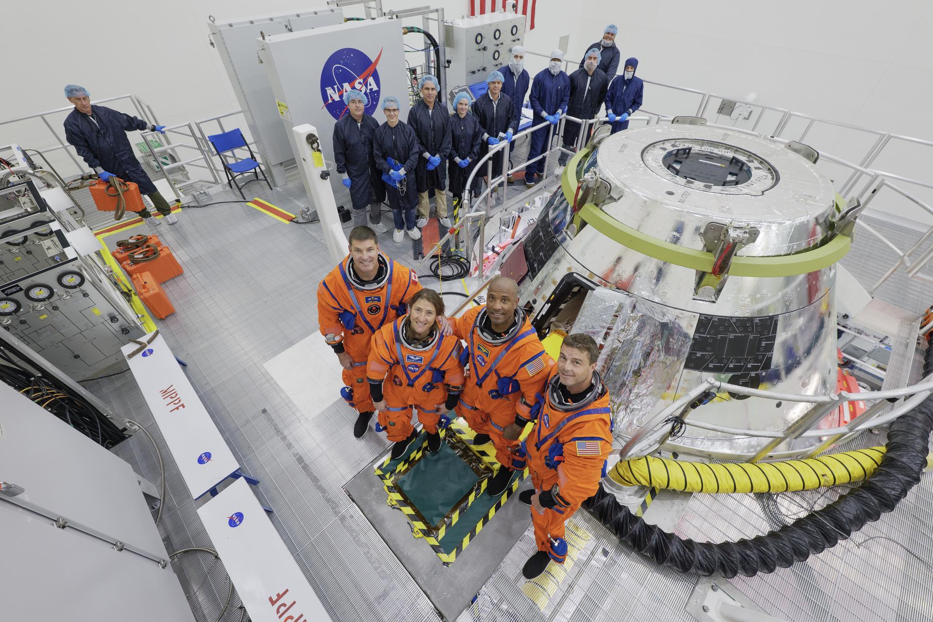 Uma fotografia de interior numa "sala limpa" da NASA, mostrando os quatro astronautas da missão Artemis II — Reid Wiseman, Victor Glover, Christina Koch e Jeremy Hansen — vestidos com fatos de voo laranjas. Eles posam junto ao módulo de tripulação da Orion, que tem um revestimento térmico prateado. Atrás deles, uma grande equipa de técnicos em uniformes azuis de proteção está de pé, ao lado de equipamento de teste com o logótipo da NASA.