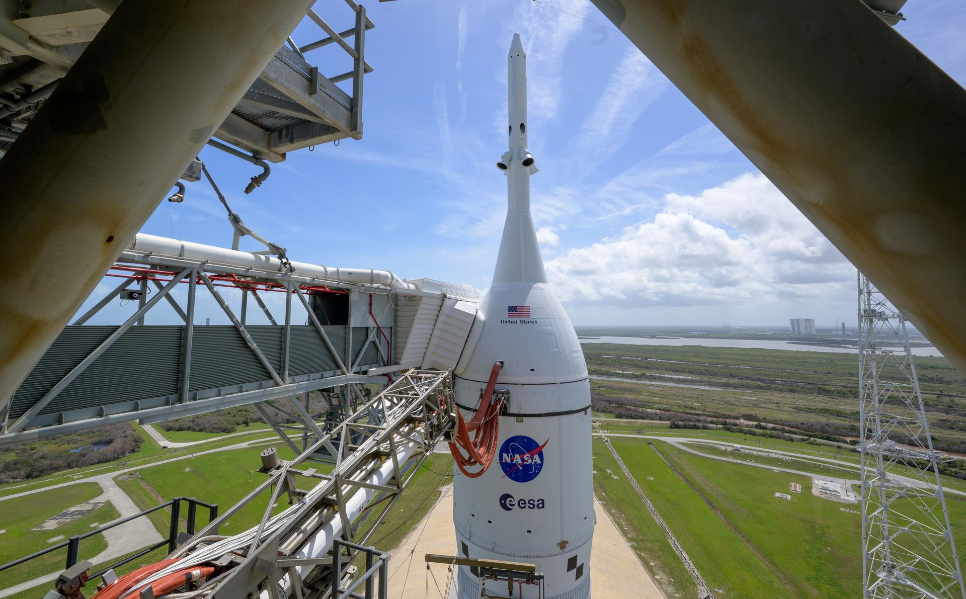 Uma vista superior e de grande angular, captada a partir da estrutura da torre de lançamento no Centro Espacial Kennedy. No centro, destaca-se a cápsula Orion e o sistema de abortagem de lançamento do foguetão SLS, exibindo as bandeiras dos EUA e os logótipos da NASA e da ESA. A nave branca está ligada por braços umbilicais e passadiços metálicos à torre de serviço. O fundo mostra a paisagem verde da Florida sob um céu azul com nuvens.