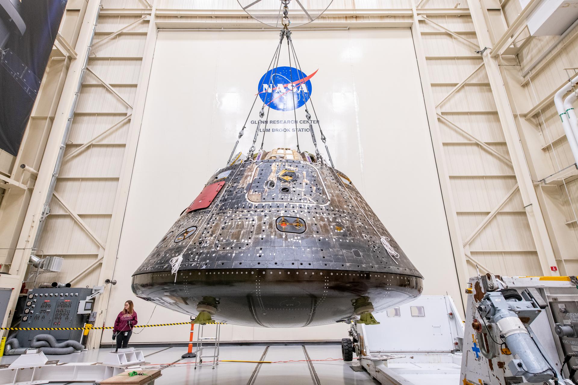 No interior de um grande hangar no Glenn Research Center da NASA, o módulo de tripulação da Orion, com um aspeto escuro e bronzeado do escudo térmico e painéis técnicos expostos, está suspenso por cabos de aço no centro. No fundo, a parede branca ostenta um grande logótipo da NASA e o texto "GLENN RESEARCH CENTER PLUM BROOK STATION". Uma técnica monitoriza a operação a partir do chão, ao lado de equipamento de suporte industrial.