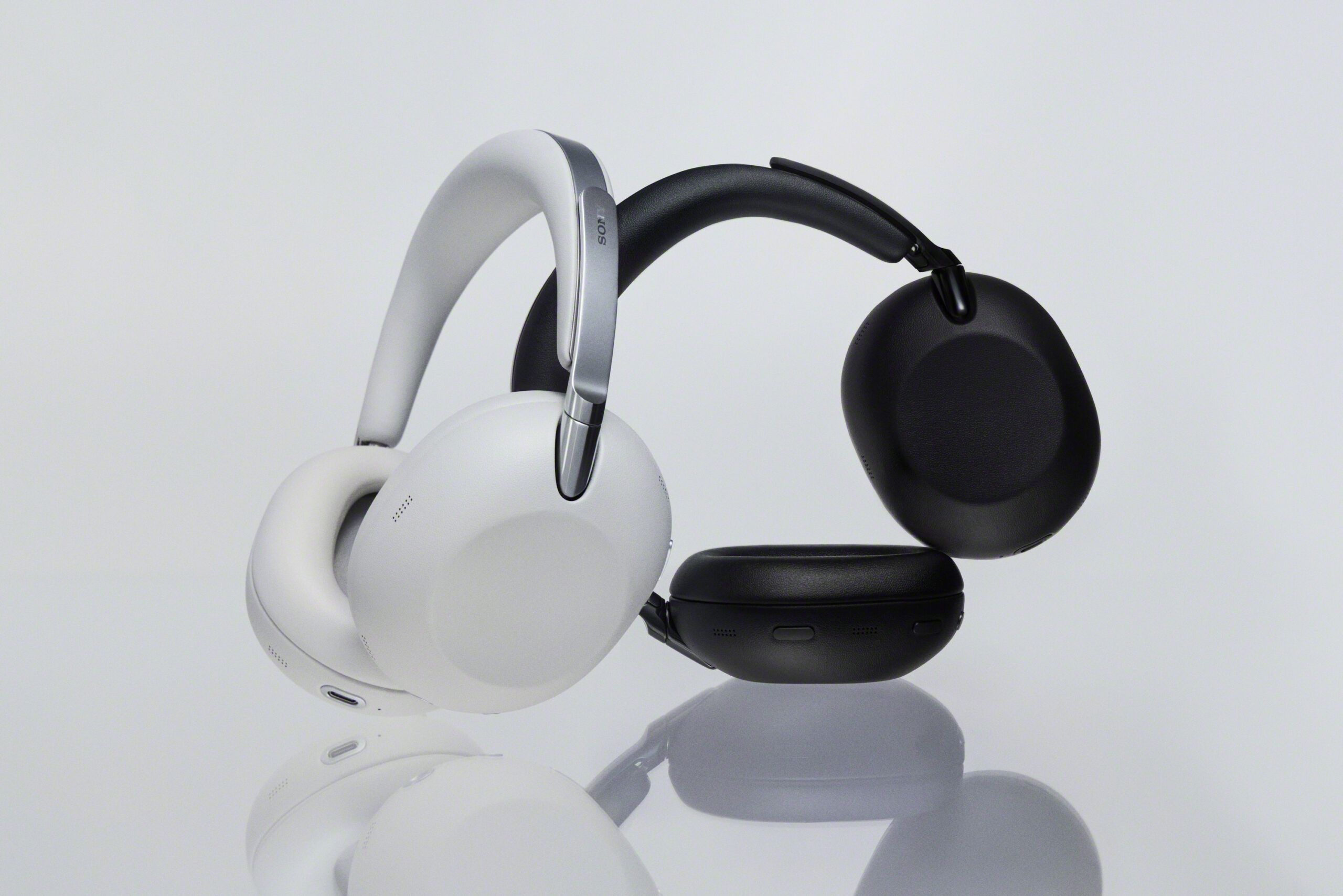 Fotografia de estúdio de dois pares de auscultadores over-ear da Sony, um num tom branco/claro com detalhes prateados e outro em preto mate. Estão pousados e encostados um ao outro sobre uma superfície branca refletora. O auscultador preto tem uma das conchas deitada, revelando os botões de controlo e as portas de ligação na sua base.