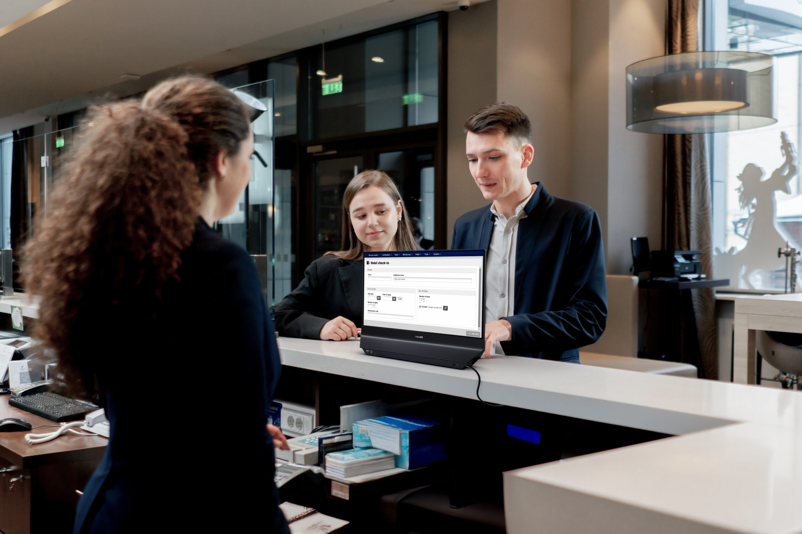 Fotografia de uma moderna recepção de hotel com uma recepcionista e dois clientes a interagir. O monitor Philips de dupla face está no balcão, com o ecrã virado para os dois clientes que olham para ele atentamente. O homem de fato está a olhar para o ecrã e a recepcionista observa, de pé atrás do balcão.