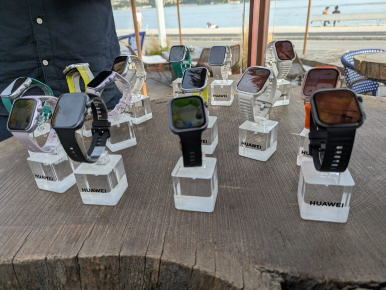 Exposição de vários smartwatches quadrados da Huawei com braceletes de diversas cores (preto, branco, verde, lilás e laranja), assentes em suportes de acrílico transparente sobre uma mesa rústica de madeira, num ambiente ao ar livre com vista para a água.