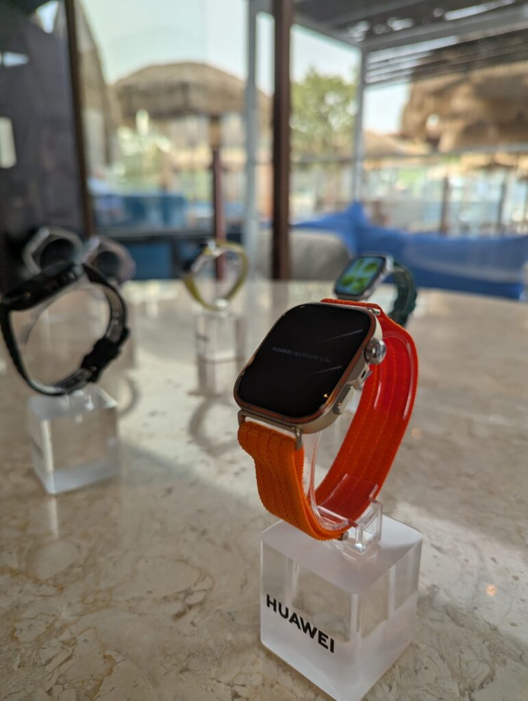 Uma fotografia em close-up de um smartwatch Huawei Watch Fit 3 Pro com uma pulseira de nylon laranja vibrante e caixa de metal prata. O relógio está em um suporte de acrílico transparente na frente, com outros smartwatches desfocados ao fundo. A cena é um interior de lounge luxuoso com vista exterior através de grandes janelas, sobre uma mesa de mármore.