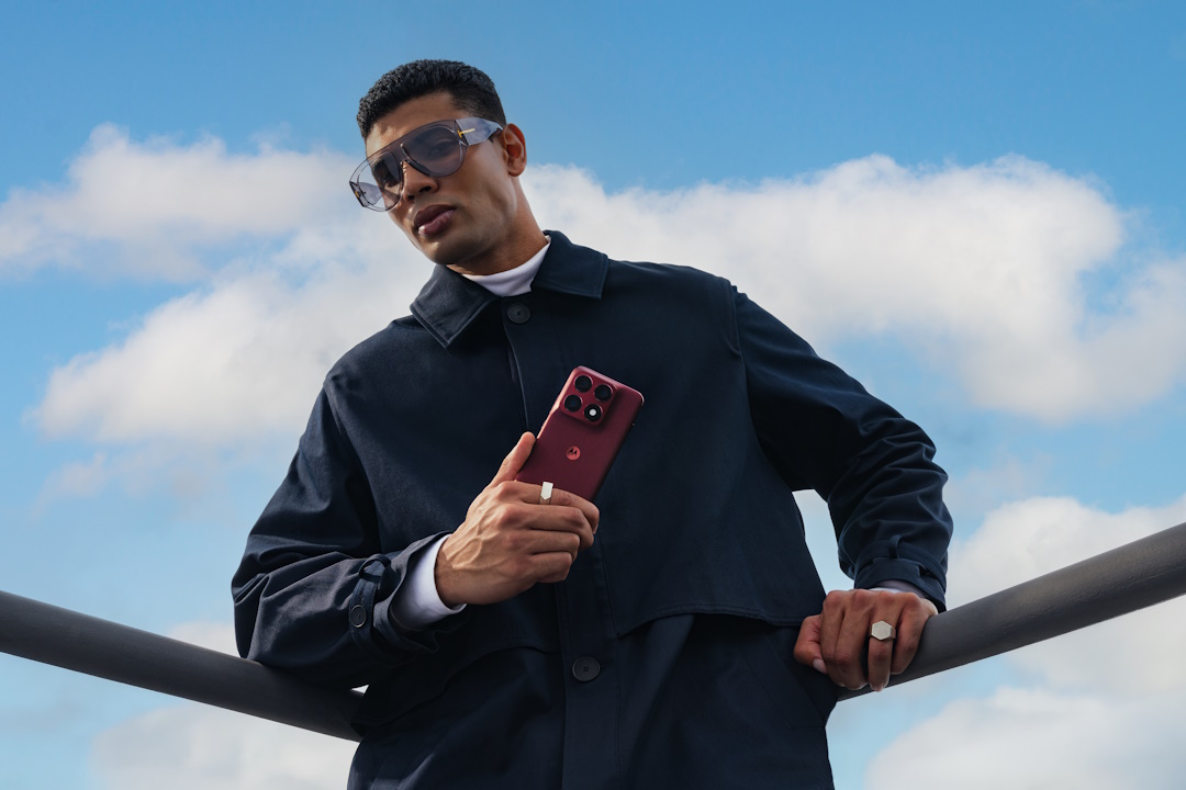 Homem elegante segura o smartphone Motorola Edge 70 Pro na cor PANTONE Zinfandel sob um céu azul com nuvens.