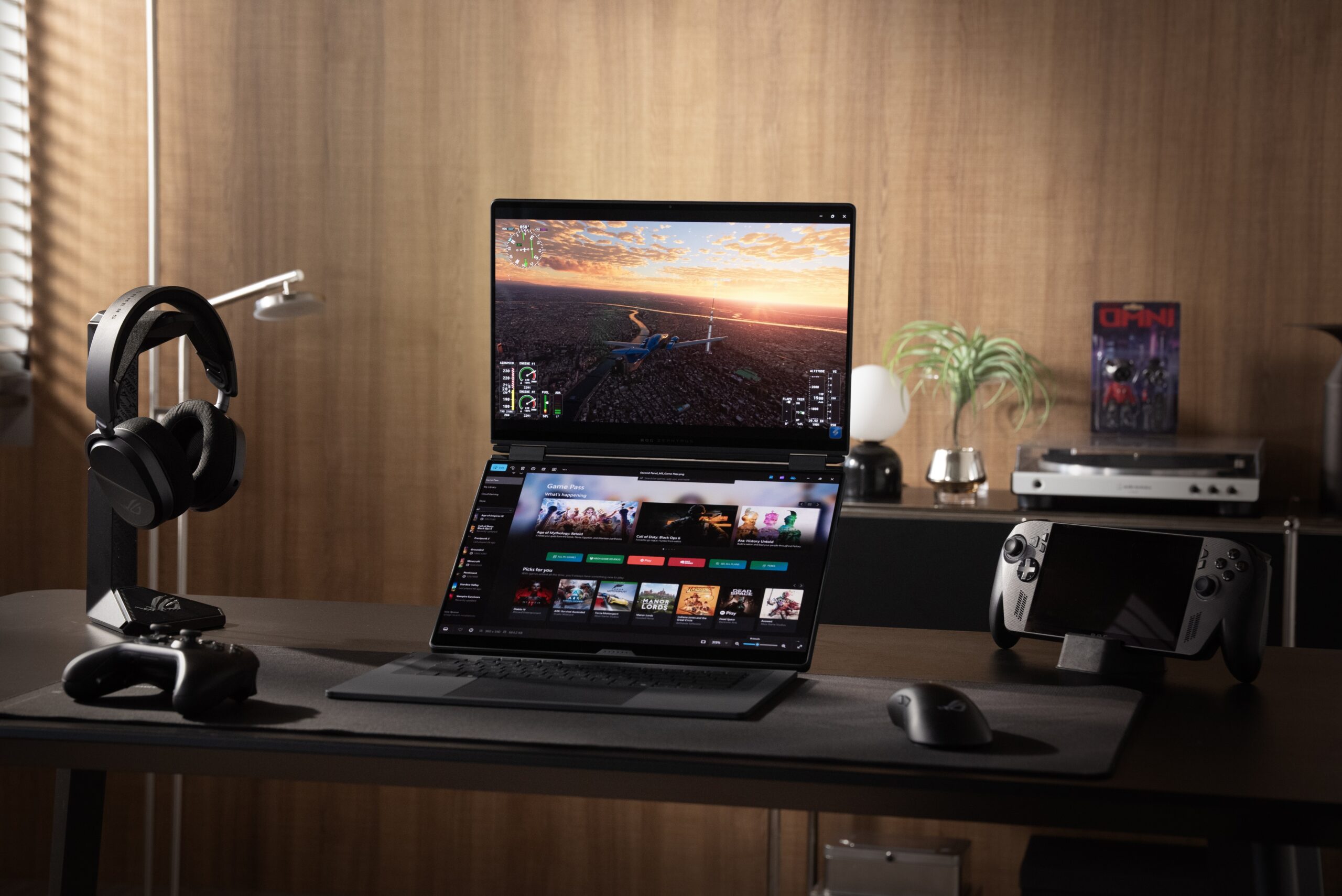 Fotografia de um setup gaming requintado numa secretária. No centro está o portátil ROG Zephyrus Duo com os seus dois ecrãs iluminados, rodeado por auscultadores num suporte, um comando, um rato e uma consola portátil ROG Ally. O fundo tem uma parede de madeira acolhedora com decoração subtil e iluminação quente.