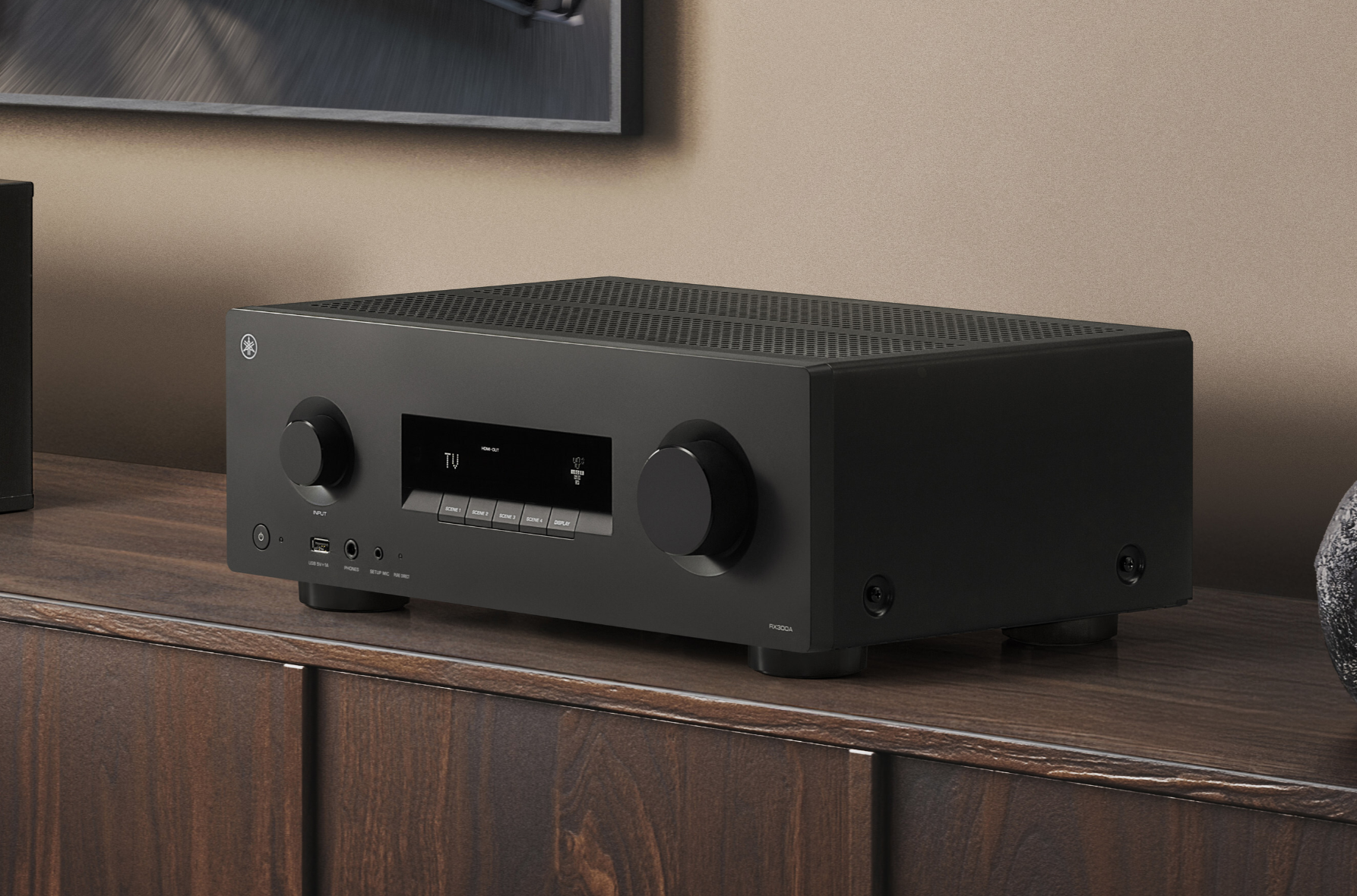 Fotografia em grande plano do receiver AV Yamaha RX300A em preto mate, assente num móvel de madeira escura. O dispositivo mostra um visor digital central com a palavra "TV", um botão de volume grande à direita e botões de controlo discretos, num ambiente de sala moderno e minimalista.
