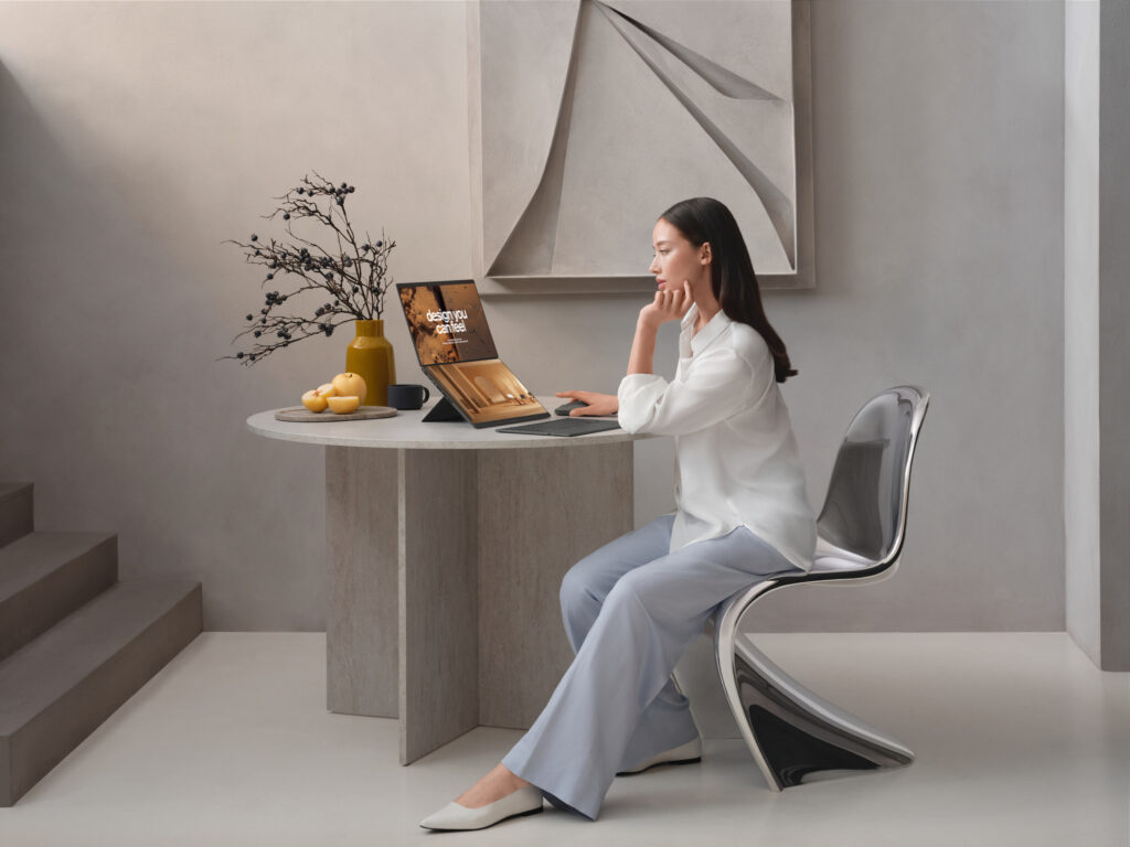 Mulher asiática sentada numa cadeira Panton cromada a uma mesa de jantar de mármore cinzento, a interagir com um portátil ASUS Zenbook Duo com dois ecrãs num ambiente de design minimalista.