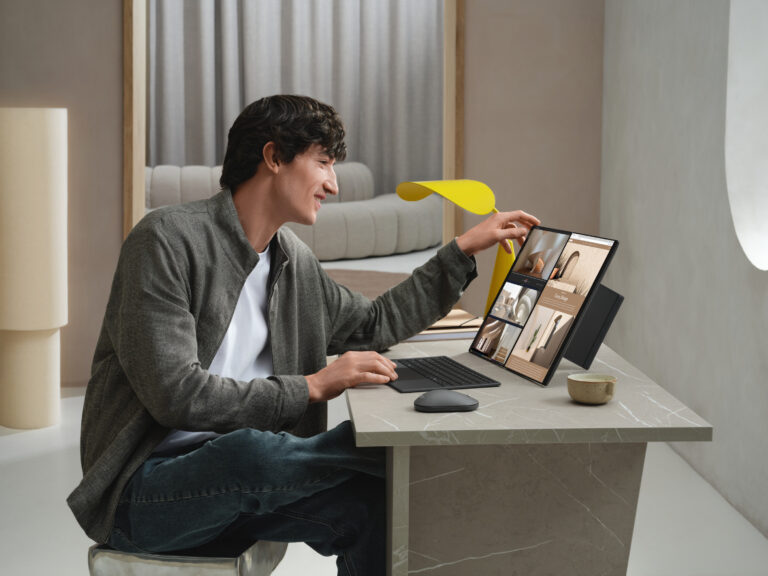 Homem jovem sentado a uma secretária moderna utiliza o portátil ASUS Zenbook DUO 2026 em modo de ecrã duplo vertical, interagindo com o ecrã tátil num ambiente de design minimalista com um candeeiro amarelo.