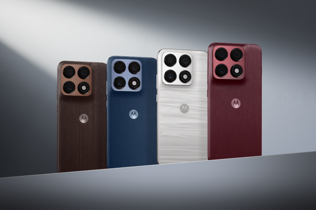 A imagem apresenta quatro unidades do novo Motorola Edge 70 Pro alinhadas lado a lado, destacando a diversidade de acabamentos da nova linha Collections™ by Motorola. Da esquerda para a direita, vemos o modelo em madeira natural (Chicory Coffee), seguido pelo azul marinho com textura de lã (Titan), um modelo branco acetinado e, por fim, a versão em seda vermelha profunda (Zinfandel). Todos os dispositivos exibem o seu generoso módulo de câmaras quádruplo e o logótipo da Motorola ao centro. A composição é iluminada por um feixe de luz diagonal que realça as texturas únicas de cada material.