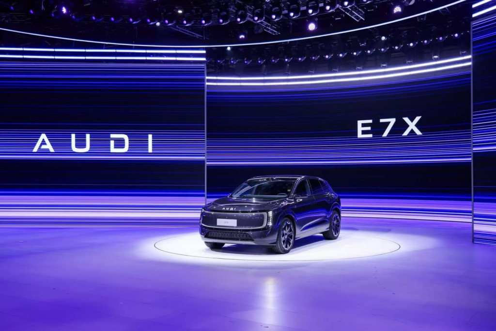 Fotografia do novo SUV elétrico Audi E7X em destaque num palco circular iluminado durante o seu lançamento mundial. O carro, de cor escura metalizada, está posicionado à frente de gigantescos painéis LED curvos que exibem linhas horizontais em tons de azul e roxo, com as inscrições "AUDI" e "E7X" em destaque. O ambiente é tecnológico e sofisticado, com iluminação de palco profissional no topo.