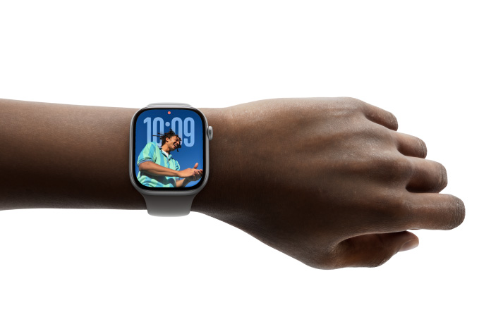 Braço de uma pessoa de pele escura a usar um Apple Watch prateado com bracelete cinzenta. O ecrã mostra as 10:09, um ponto vermelho de notificação e a foto de um jovem a sorrir com uma camisa às riscas contra um céu azul. Fundo totalmente branco.