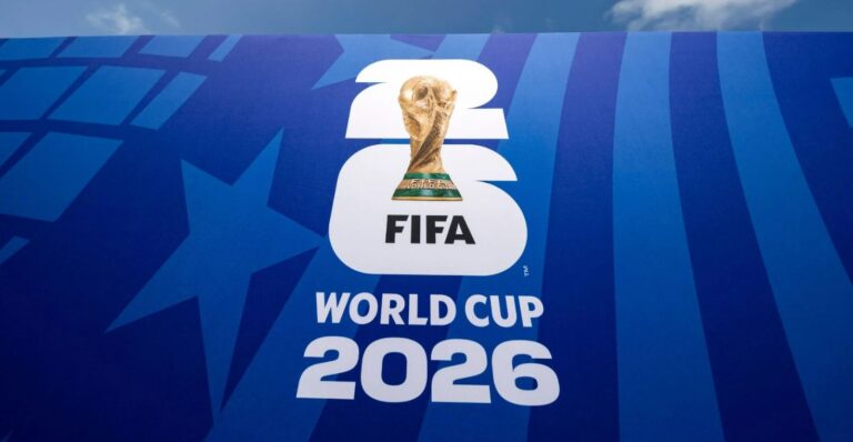Painel azul vibrante com o logótipo oficial do Campeonato do Mundo da FIFA 2026. O design destaca os algarismos "26" empilhados em branco, com a icónica taça dourada do Mundial posicionada no centro do número seis, acompanhada da inscrição "WORLD CUP 2026" na base.