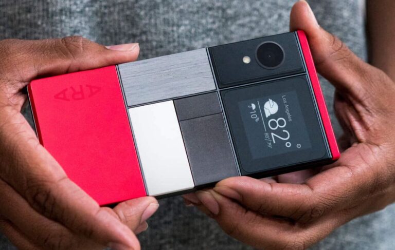 Fotografia de perto das mãos de uma pessoa a segurar o protótipo do smartphone modular Project Ara, destacando a sua traseira composta por blocos físicos intercambiáveis de várias cores, um módulo de câmara fotográfica e um mini-ecrã a exibir a meteorologia.