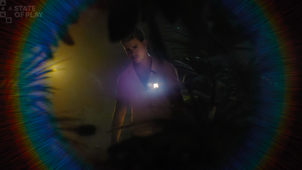 Um jovem de camisa com uma luz LED brilhante no peito é emoldurado por um círculo de visão em escuridão profunda, cercado por distorção óptica em arco-íris. O fundo mostra folhagem tropical e uma luz ambiente difusa. O logotipo "PLAYSTATION | STATE OF PLAY" está no canto superior esquerdo. Esta é uma cena do arquivo "until down 2 playstation megaverso.jpg".