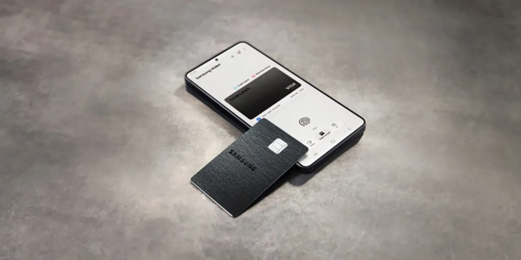 Cartão de crédito físico preto da Samsung apoiado sobre um smartphone que tem o ecrã ligado a exibir a aplicação Samsung Wallet, tudo sobre uma superfície cinzenta texturizada.