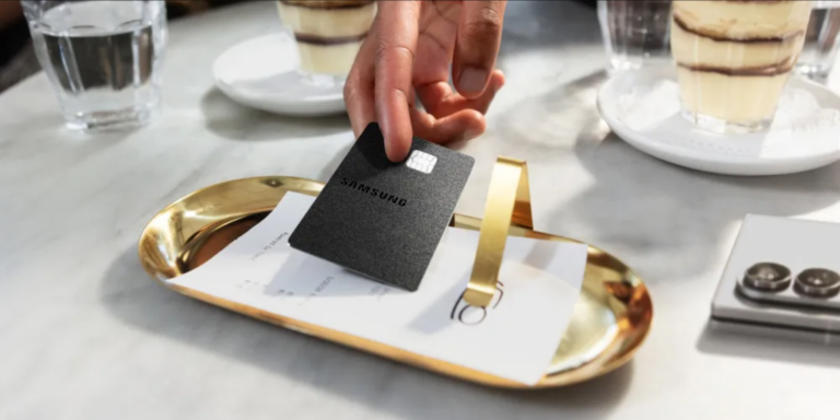 A mão de uma pessoa a colocar um cartão de crédito Samsung Galaxy Card em metal preto sobre um tabuleiro valet dourado numa mesa de mármore.